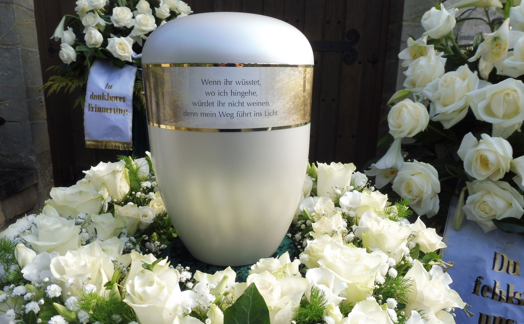 Detailaufnahme einer weißen Urne mit goldener Verzierung und einem eingravierten Spruch, umgeben von weißen Rosen und Trauerschleifen.