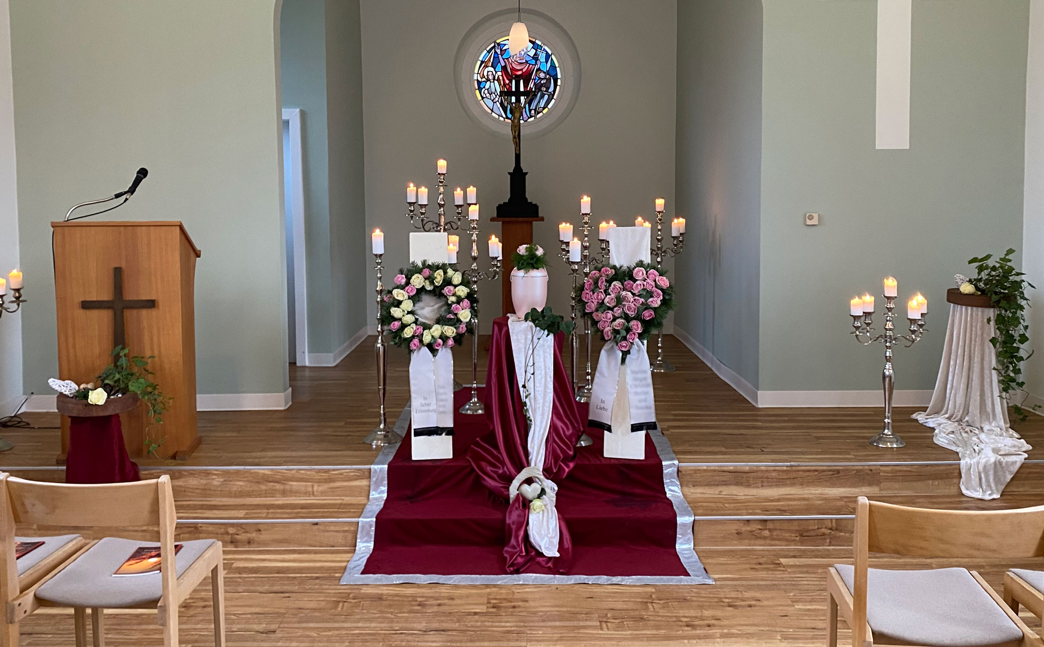 Stilvolle Trauerfeier mit Urne, Kerzenständern und Blumen in einer hellen Kapelle mit Holzboden und Altar mit Christusfigur im Hintergrund.