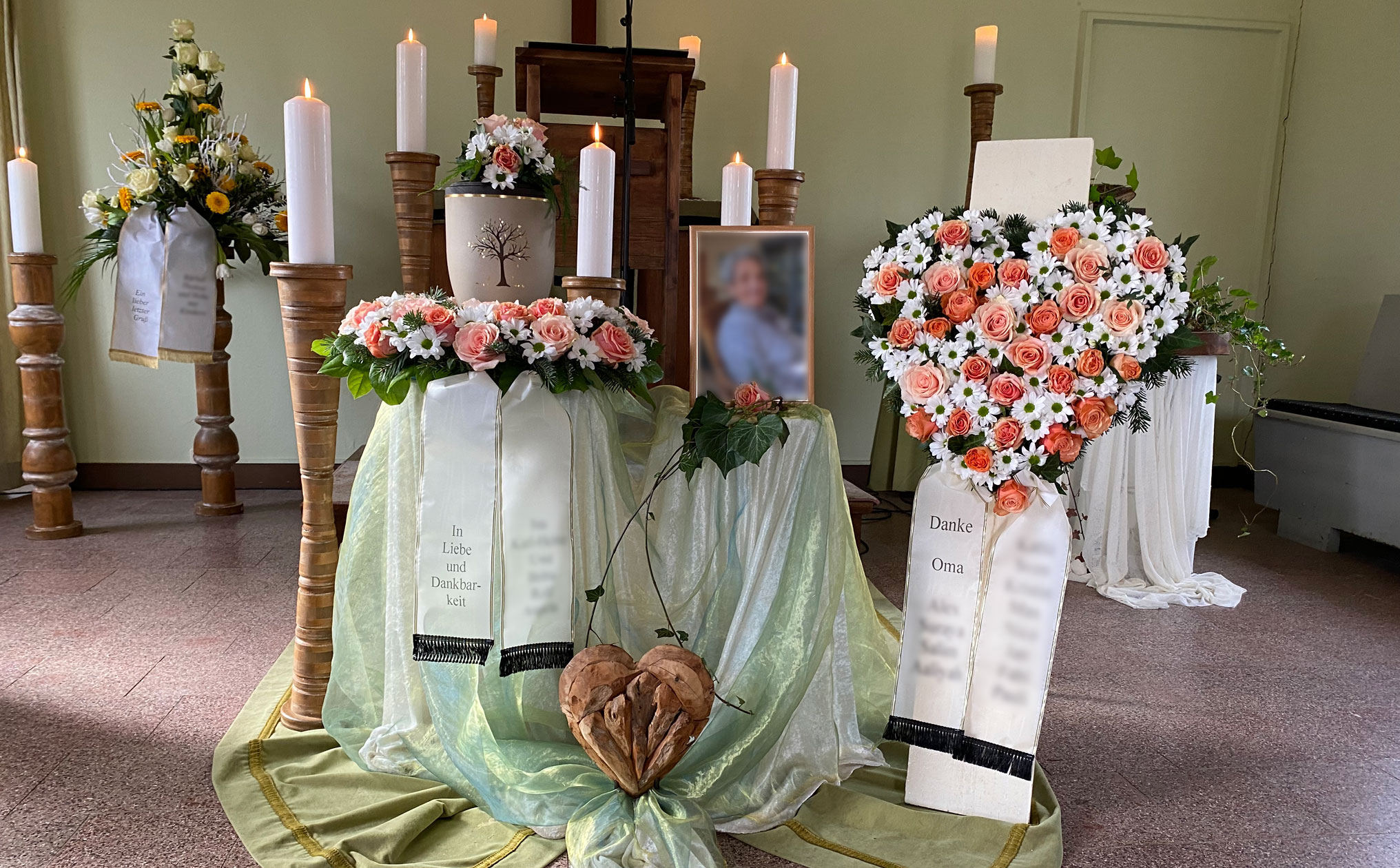 Liebevoll gestaltete Urnenabschiednahme mit Kerzen, Blumengestecken und einer Urne mit Baum-Motiv, umgeben von Trauerschleifen mit persönlichen Botschaften.