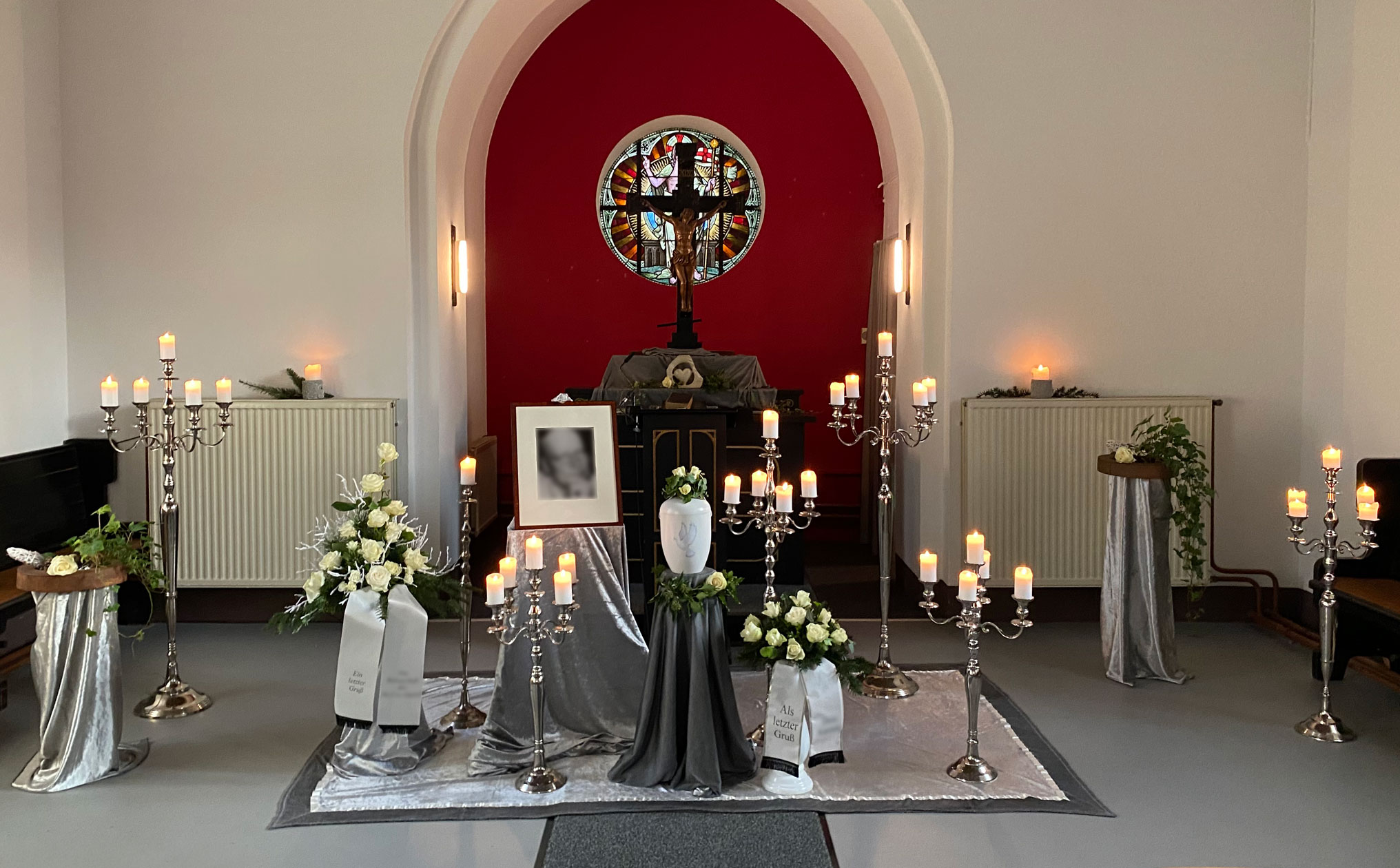 Stimmungsvolle Trauerfeier in einer Kapelle mit rotem Altarbereich, zahlreichen Kerzenleuchtern, einer Urne und weißen Blumen als Abschiedsgesteck.